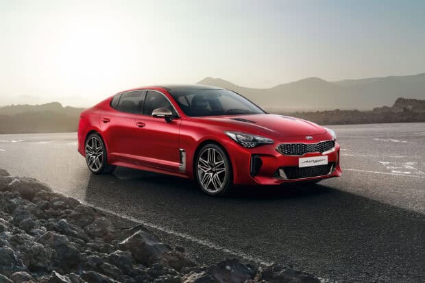 A red Kia Stinger is parked on an open mountain road with rocky terrain and hills in the background, 2K Desktop Wallpaper