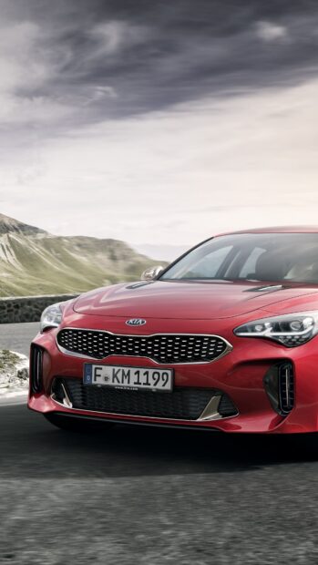 A red Kia car is driving smoothly on a mountain road under cloudy skies with scenic hills, 4K Mobile Wallpaper