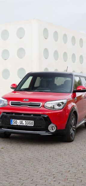 A red Kia car is parked near a white building with circular windows in the background, 2K Mobile Wallpaper
