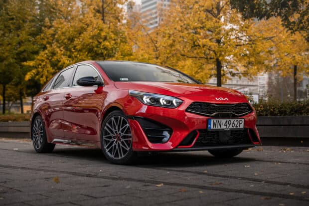 A red Kia car is parked on a city street surrounded by autumn trees with yellow leaves, 2K Desktop Wallpaper