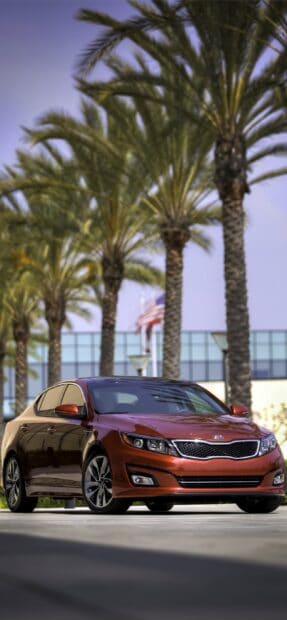A red Kia car is parked outdoors with palm trees and a waving American flag in the background, 2K Mobile Wallpaper