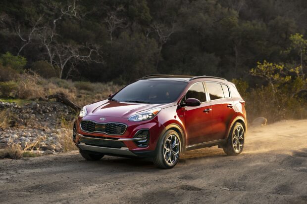 A red Kia car is driving on a dirt road surrounded by trees and rocks in a natural setting, 2K Desktop Wallpaper