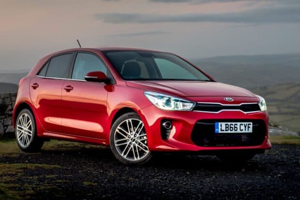 A red Kia model is parked on a gravel area with a scenic countryside view in the background featuring hills and cloudy skies, HD Desktop Wallpaper