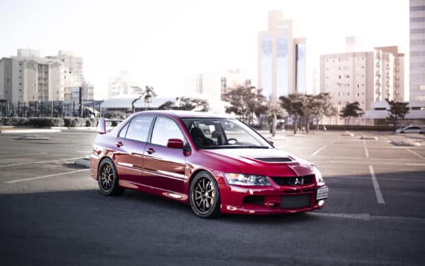 A red Lancer Evo is parked in an empty urban lot with buildings in the background, 2K Desktop Wallpaper