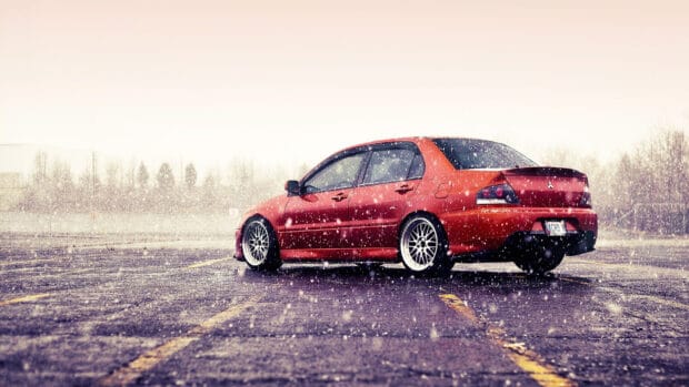 A red Lancer Evo is parked in a snowy parking lot with visible snowflakes falling around the car, HD Desktop Wallpaper