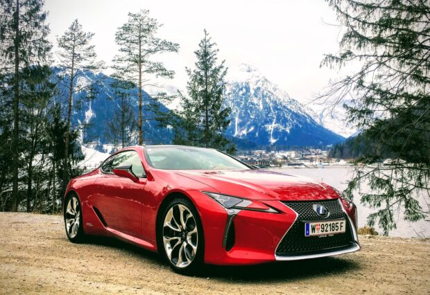 A red Lexus LC is parked on a dirt road near a lake with snow covered mountains in the background, 2K Desktop Wallpaper
