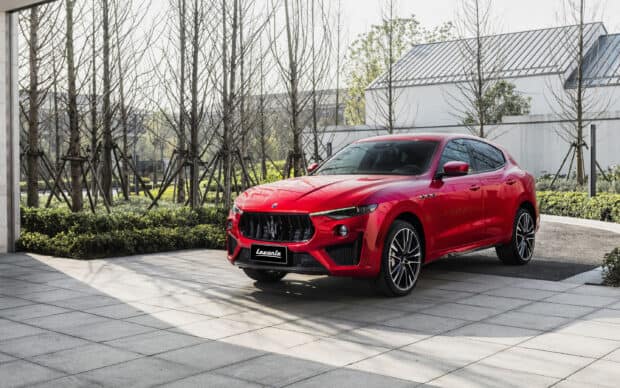 A red Maserati Levante is parked near tiled pavement with trees and modern buildings in the background, 2K Desktop Wallpaper