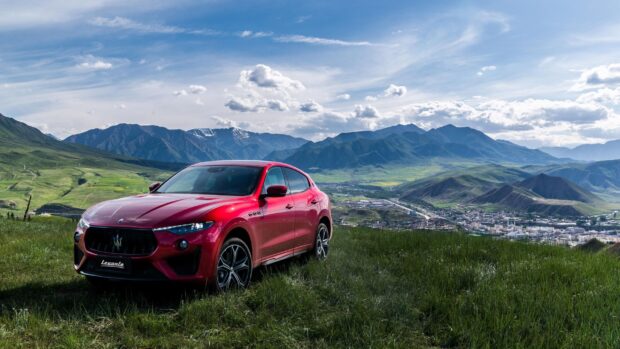 A red Maserati Levante is parked on a grassy hill with mountains and a town in the background, 2K Desktop Wallpaper