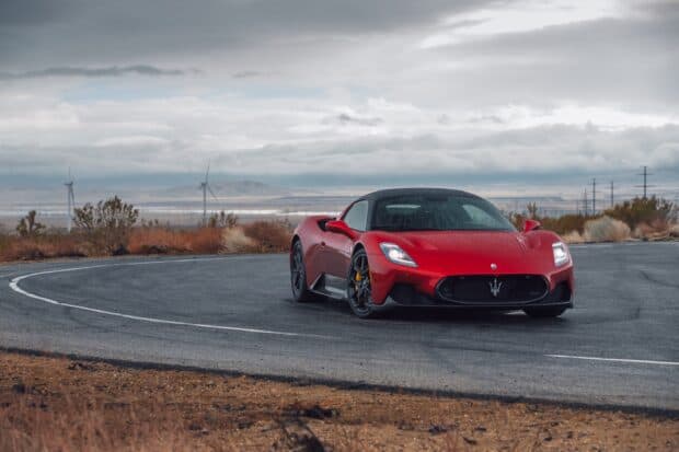 A red Maserati MC20 sports car is parked on a wet curved road with wind turbines and cloudy sky in the background, 2K Desktop Wallpaper