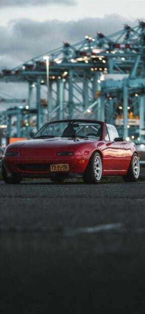 A red Mazda MX 5 Miata is parked on the road with large industrial cranes in the background, 2K Phone Wallpaper