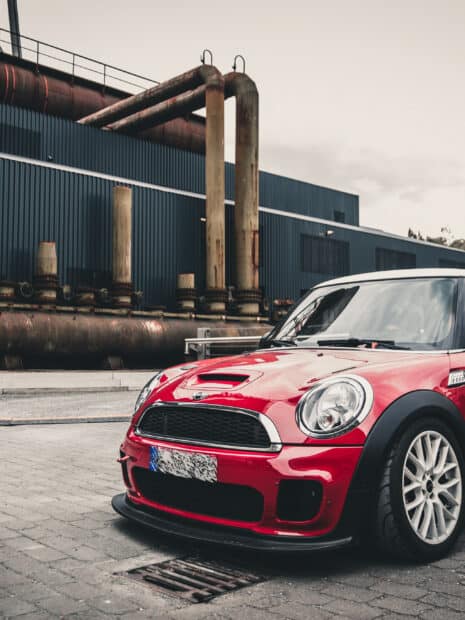 A red Mini Cooper is parked by rusty industrial pipes outside a building, 2K Mobile Wallpaper