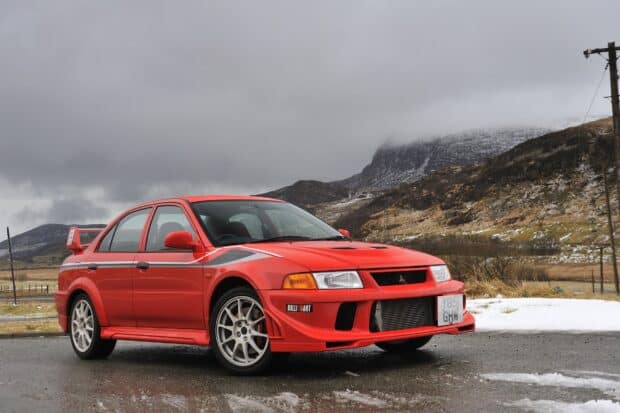 A red Mitsubishi Lancer Evo stands on a wet mountain road with cloudy sky and snowy hills, HD Desktop Wallpaper