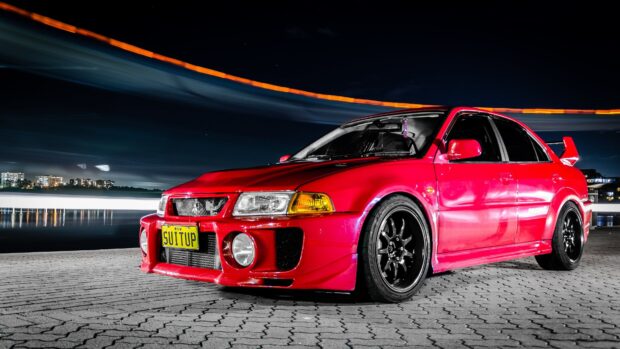 A red Mitsubishi Lancer Evo is parked on a tiled area with city lights in the background, 4K Desktop Wallpaper