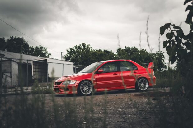 A red Mitsubishi Lancer Evo is parked on a gravel surface with trees and a cloudy sky in the background, HD Desktop Wallpaper