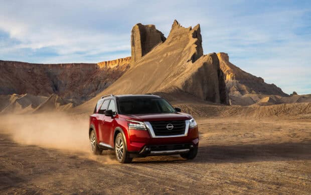 A red Nissan Pathfinder is driving through dusty desert terrain with rock formations in the background, HD Desktop Wallpaper