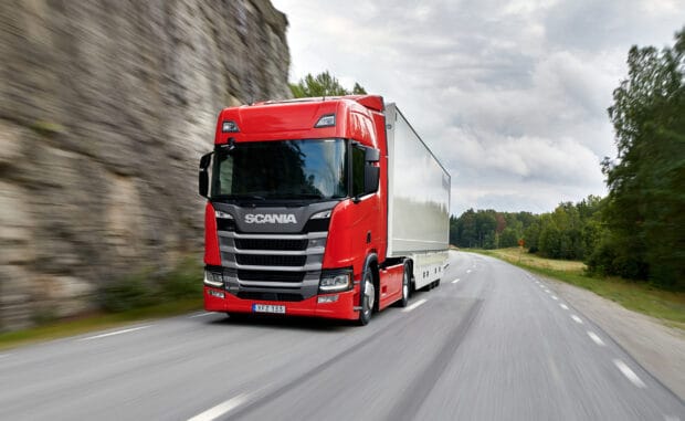 A red Scania truck drives fast on a winding road surrounded by trees and cliffs, 2K Desktop Wallpaper