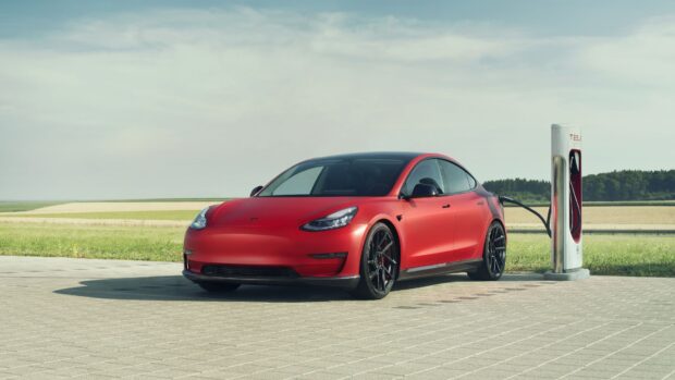 A red Tesla Model 3 is charging outdoors on a sunny day with fields in the background, 4K Desktop Wallpaper