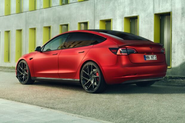 A red Tesla Model 3 is parked beside a contemporary building with yellow window frames in the background, HD Desktop Wallpaper