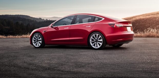 A red Tesla Model 3 is parked on a road with hills and trees in the background, 2K Desktop Wallpaper