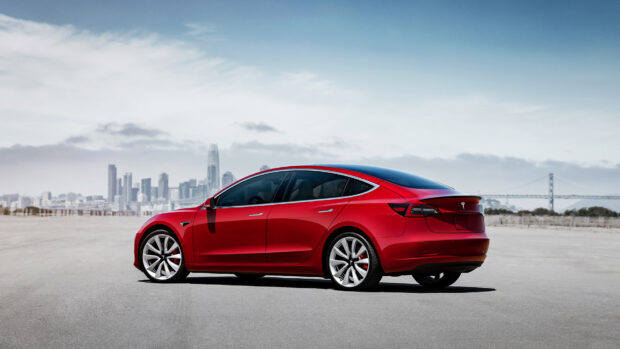 A red Tesla Model 3 is parked on a wide road with a city skyline and bridge in the background, HD Desktop Wallpaper