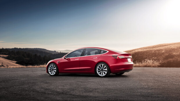 A red Tesla Model 3 is parked on a paved road with hills and a clear sky in the background, HD Desktop Wallpaper