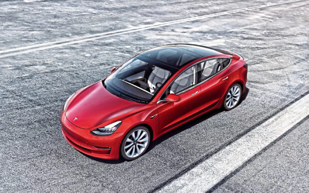 A red Tesla Model 3 is parked on a textured racetrack surface under sunlight, 2K Desktop Wallpaper