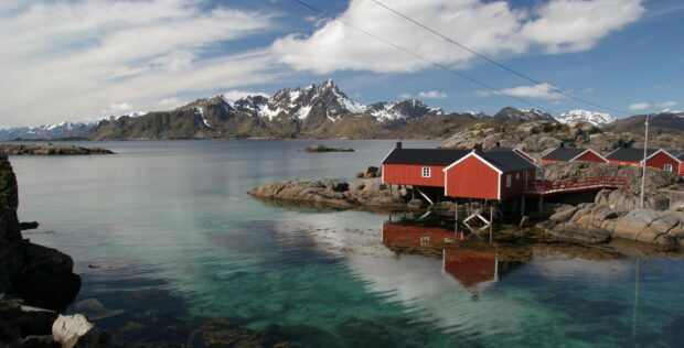 The Norway Wallpapers show red houses standing on rocky shores with clear water reflecting snowy mountains in the background, 2K Desktop Wallpaper