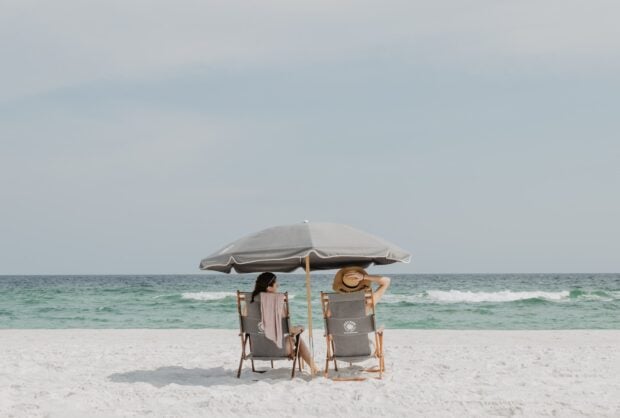 Two people sit under a beach umbrella facing the ocean on a sandy shore, HD Desktop Wallpaper