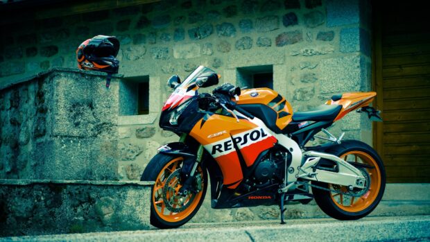 A bright orange Repsol Honda street bike is parked near a stone wall with a matching helmet placed on a ledge, 4K Desktop Wallpaper