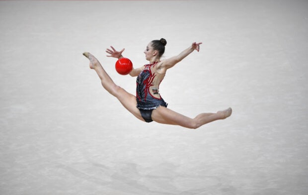 A rhythmic gymnastics athlete performs a split leap while holding a red ball in a competition, HD Desktop Wallpaper