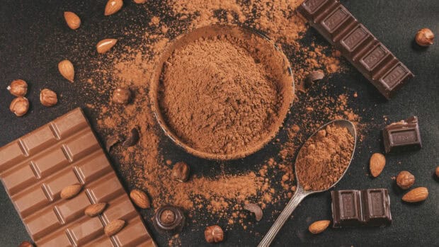 A bowl of rich cocoa powder surrounded by chocolate bars and nuts on a dark surface featuring Cocao Wallpapers, 4K Desktop Wallpaper