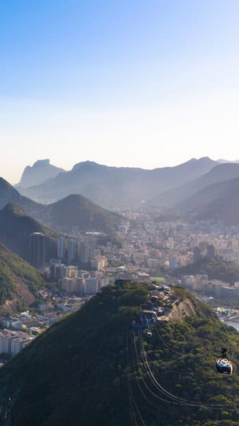 A cable car travels over green hills with Rio De Janeiro Wallpapers cityscape in the background, 2K Mobile Wallpaper