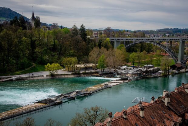 The River Aare flows peacefully under a historic bridge with surrounding greenery in Bern Switzerland, HD Desktop Wallpaper