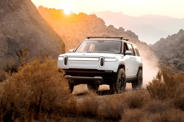 A white Rivian automotive driving through a dusty desert road with rocky mountains in the background, 2K Desktop Wallpaper