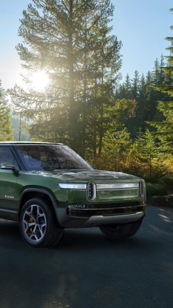 A green Rivian Automotive SUV is parked on a forest road with sunlight shining through the trees, 4K Phone Wallpaper