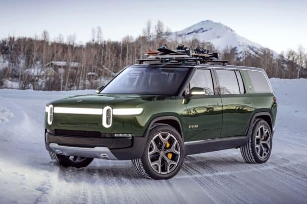 A Rivian automotive vehicle is parked on a snow covered road near trees and a mountain, 2K Desktop Wallpaper