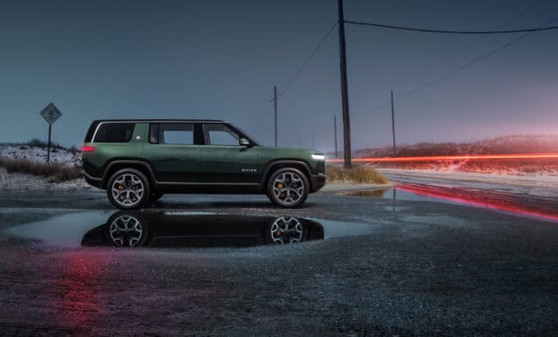 A Rivian Automotive vehicle is parked next to a puddle reflecting its image on a quiet road at dusk, HD Desktop Wallpaper