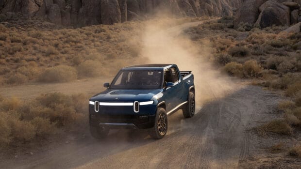 A Rivian electric truck drives through a dusty desert road surrounded by rocky terrain, HD Desktop Wallpaper