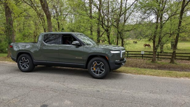 A Rivian Automotive vehicle is parked on a road next to a lush green field with trees and horses in the background, HD Desktop Wallpaper