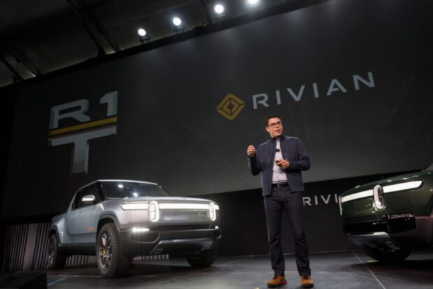 A man presenting Rivian electric trucks on stage with Rivian Automotive Wallpapers branding, 2K Desktop Wallpaper
