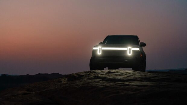 A Rivian Automotive vehicle with its distinctive front lights glowing at dusk on rocky terrain, 2K Desktop Wallpaper