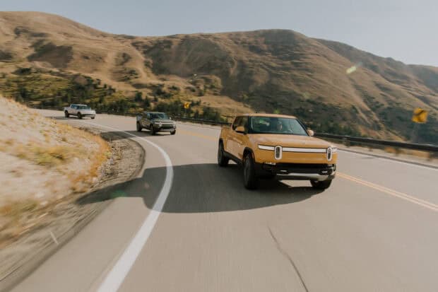 A group of Rivian vehicles is driving along a winding mountain road under clear skies, 2K Desktop Wallpaper