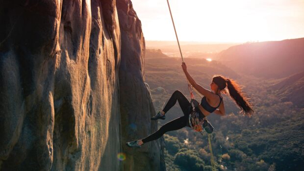 A woman enjoys rock climbing on a cliff with sunlight illuminating the scene, 4K Desktop Wallpaper