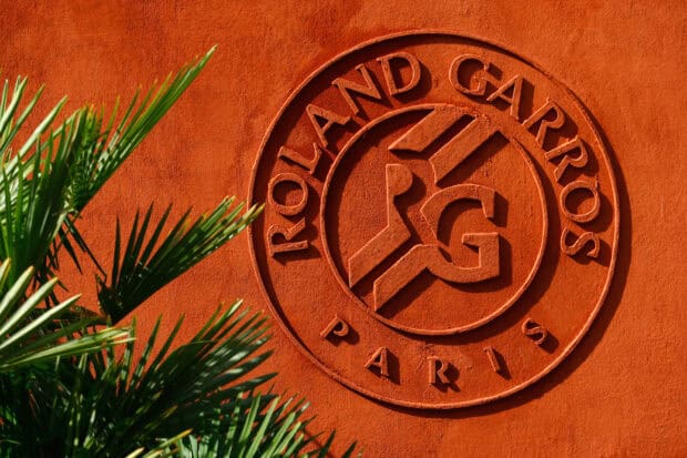 The French Open Roland Garros logo is engraved on a clay wall with green palm leaves in front, HD Desktop Wallpaper
