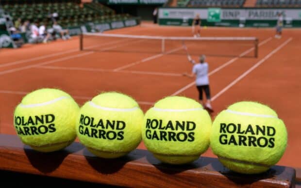 Four Roland Garros tennis balls rest on a wooden railing with players in the background on a clay court, 2K Desktop Wallpaper