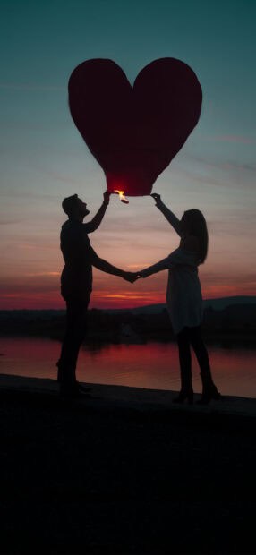 A love couple is holding a heart shaped lantern at sunset by the water, HD Phone Wallpaper
