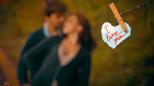 A blurred love couple standing in the background with a heart shaped note saying I love you hanging on a branch, 4K Desktop Wallpaper