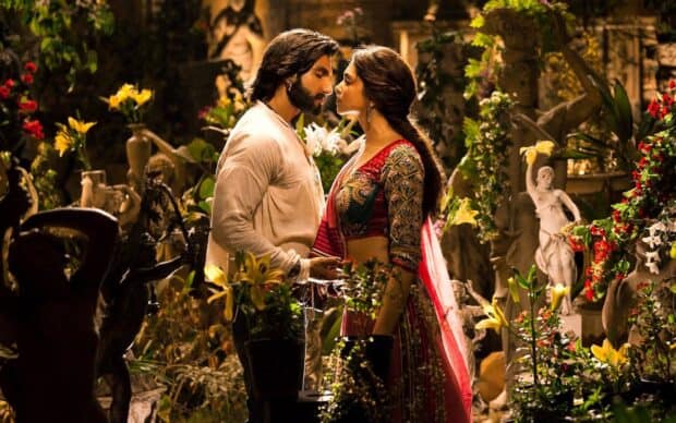 A couple shares an intimate moment surrounded by flowers and statues in a Bollywood setting, HD Desktop Wallpaper
