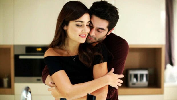 A man lovingly embraces a smiling woman in a cozy kitchen setting featuring Hande Ercel wallpapers, HD Desktop Wallpaper