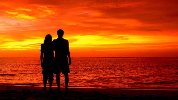 A love couple stands together watching a beautiful orange sunset by the sea, 4K Desktop Wallpaper